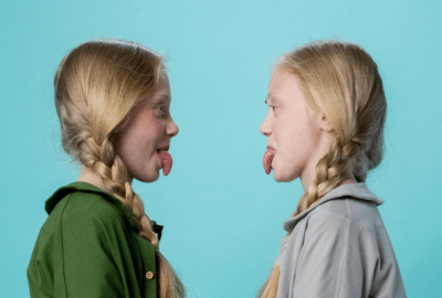 Identical twin girls with blonde hair look at each other, sticking their tongues out.