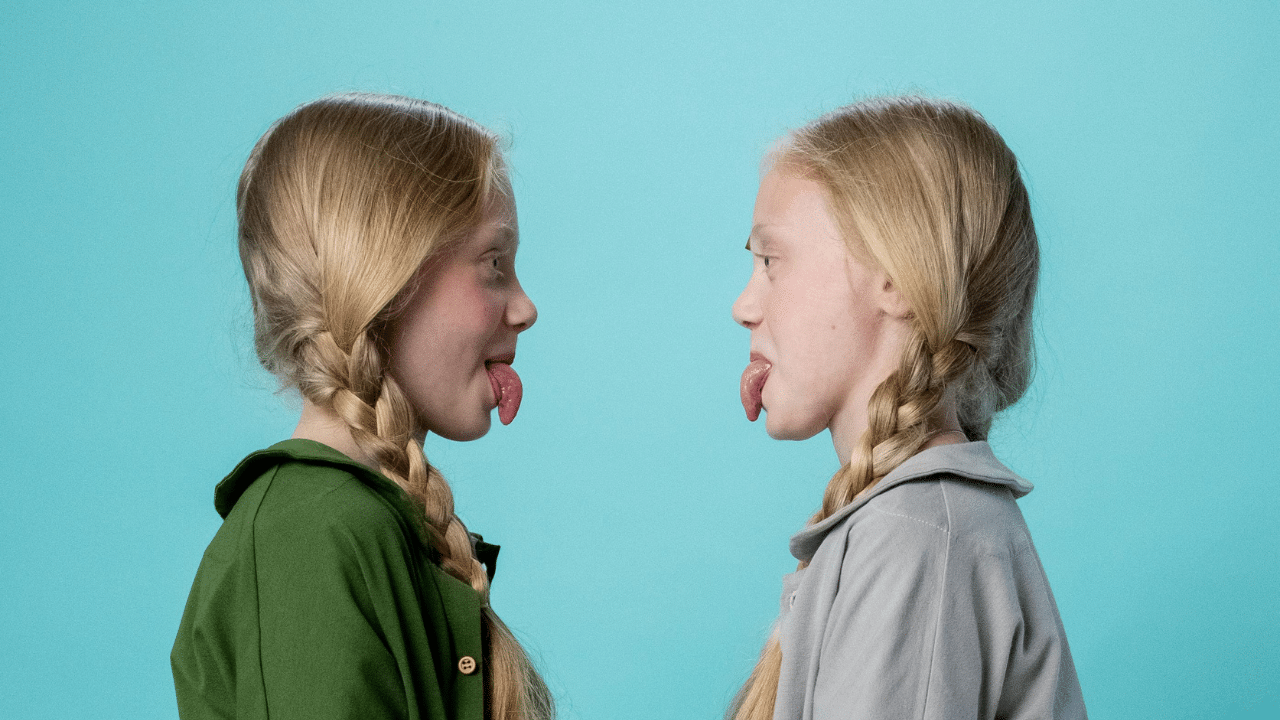 Identical twin girls with blonde hair look at each other, sticking their tongues out.