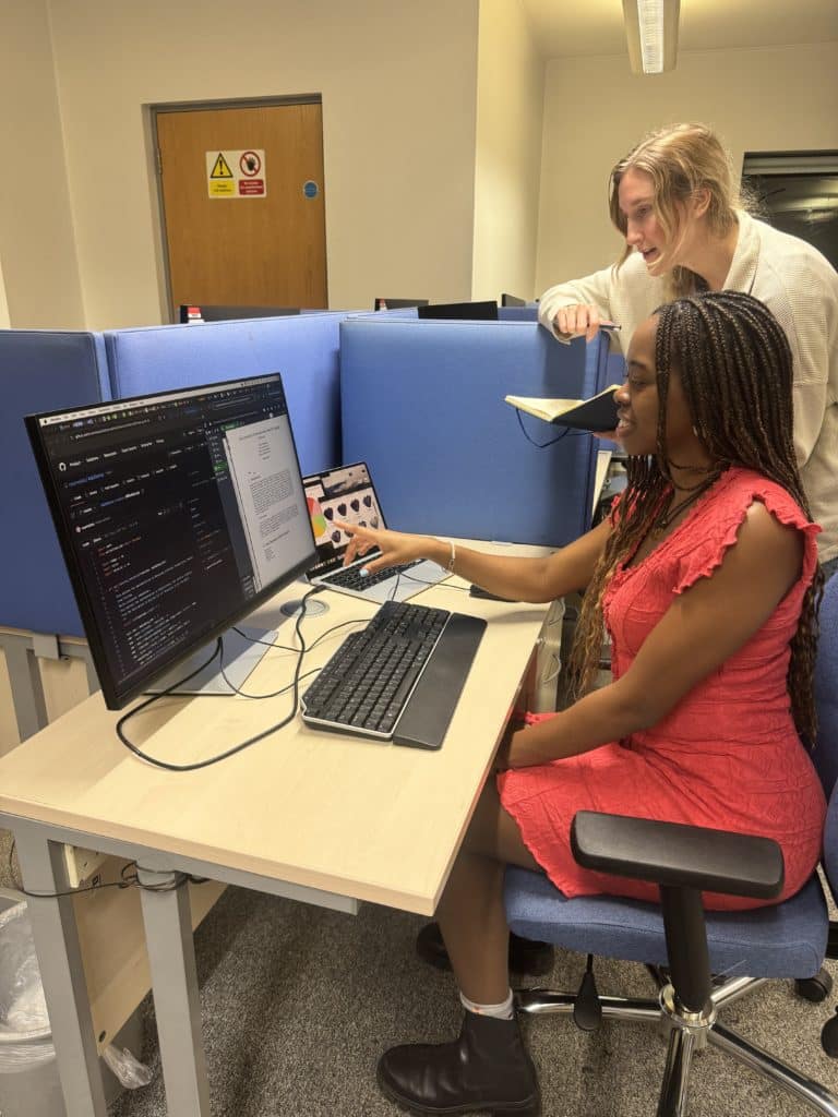 Emma sitting at a computer, pointing at the screen, showing another PhD student her research