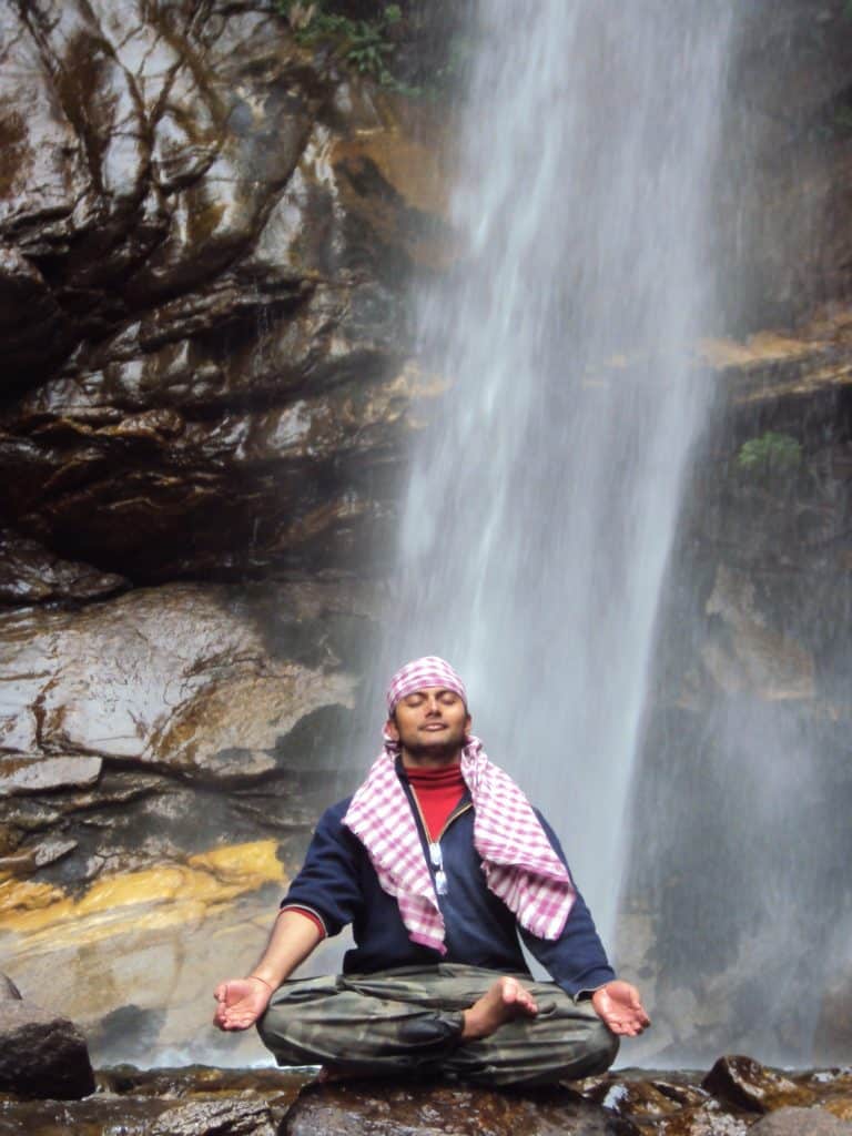 Nishant sitting cross-legged in front of a waterfall, meditating.