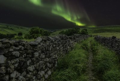 The Aurora Borealis, or Northern Lights, above a stone wall and green fields. The Aurora appears as a green curved streak in the night sky.