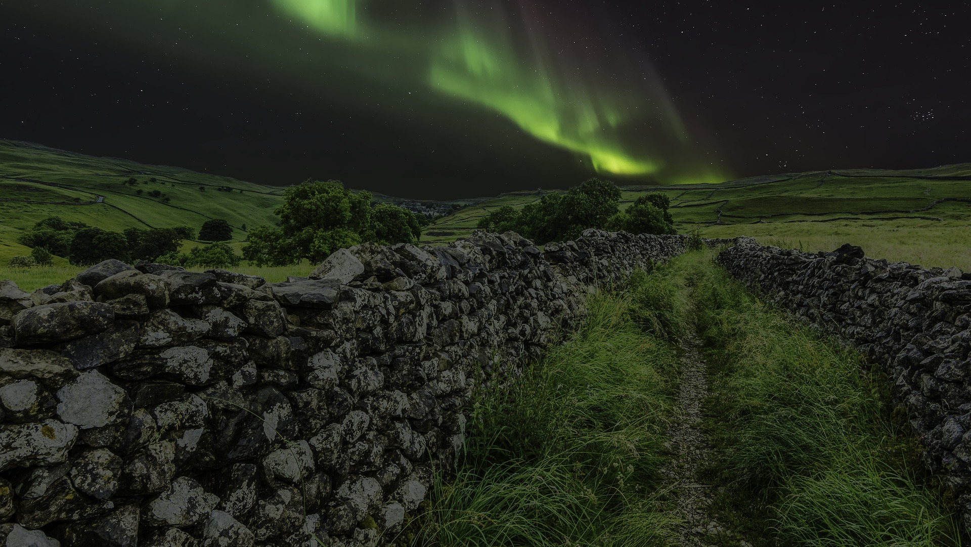 The Aurora Borealis, or Northern Lights, above a stone wall and green fields. The Aurora appears as a green curved streak in the night sky.